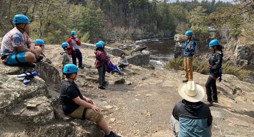A group of young people wearing safety gear listen to an instructor while preparing to rock climb. There is a river in the background. 